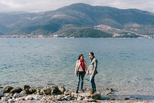 Man and woman are holding hands on the stones by the sea against the background of mountains. High quality photo