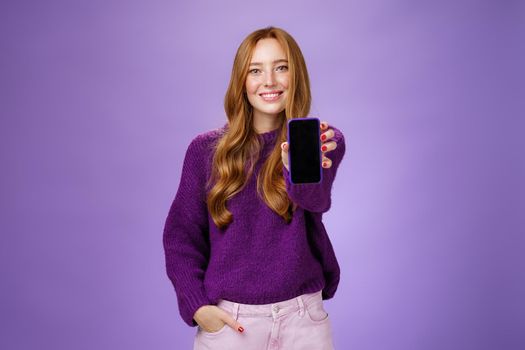 Girl shows smartphone screen at camera to ask opinion of friend smiling broadly with optimistic and joyful expression holding hand in pocket promoting mobile phone or app over purple background.