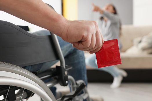 Close-up of man sit in wheelchair hold package of condom, woman laugh on couch pointing with finger. Disabled people, intimate life with disability concept