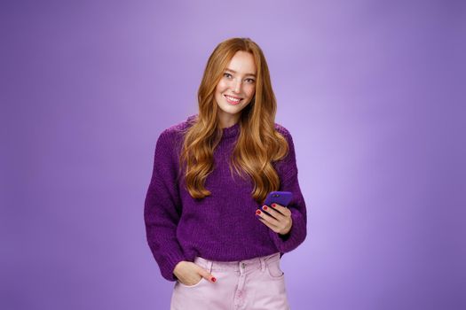 Friendly-looking relaxed and joyful nice 20s female student writing post in social network using mobile phone holding one hand in pocket and texting friend with smartphone, smiling satisfied.