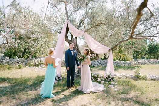 Groom stands with bride near a tree in a green grove in front of the wedding ceremony master. High quality photo