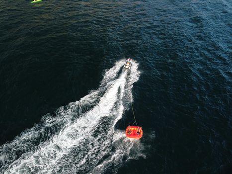 Happy people swim on air mattress behind a high speed water bike. Tourists ride on inflatable watercraft boat. Sea attraction. Water boat skating people on large Inflatable raft. Aerial copter view