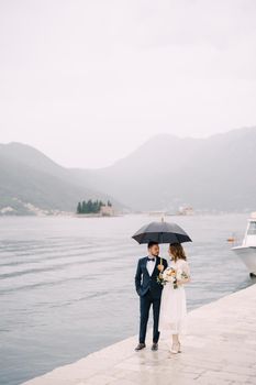 Bride and groom with an umbrella stand on the pier by the sea against the background of mountains. High quality photo