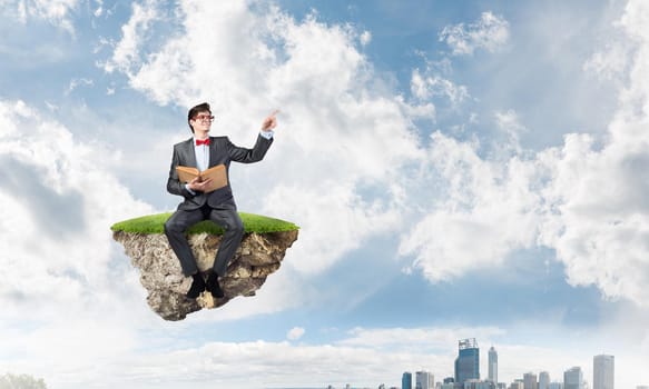 Young student with a book sits on a flying island. The cityscape in the background
