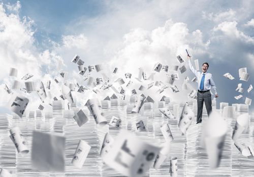 Businessman keeping hand with book up while standing among flying papers with cloudly skyscape on background. Mixed media.
