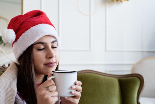 Merry Christmas and Happy New Year. Young brunette woman in santa hat and cozy plaid sitting on green couch alone in a decorated for Christmas living room, having cup of hot drink