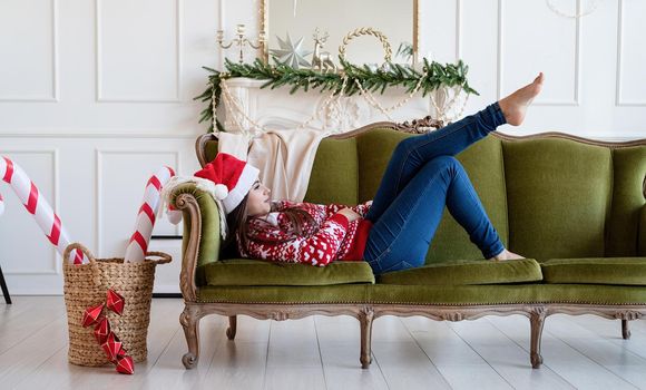 Merry Christmas and Happy New Year. Young brunette woman in santa hat lying on green couch alone in a decorated for Christmas living room