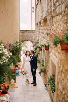 Bride and groom walk along a narrow old street decorated with blooming flowerpots. High quality photo
