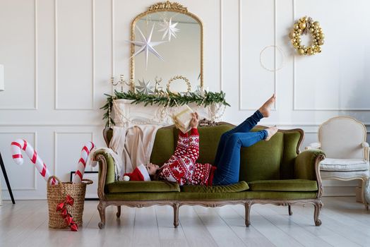 Merry Christmas and Happy New Year. Young brunette woman in santa hat lying on green couch alone in a decorated for Christmas living room