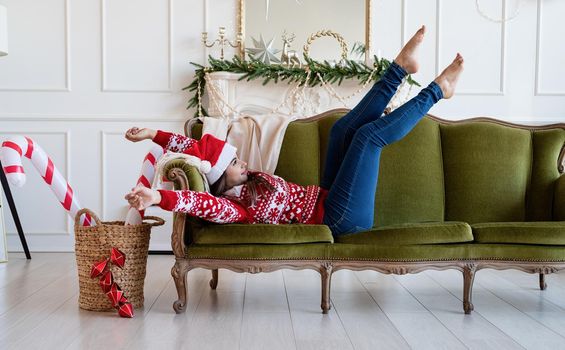 Merry Christmas and Happy New Year. Young brunette woman in santa hat lying on green couch alone in a decorated for Christmas living room