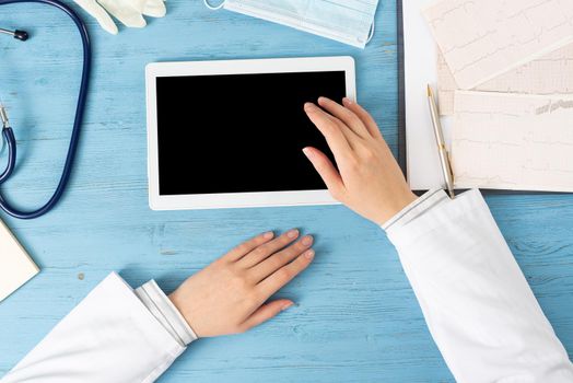 Top view of doctor hands with tablet computer. Cardiac examination in hospital. Therapist sitting at blue wooden desk with stethoscope, medical gloves and mask. Examination and consultation in clinic