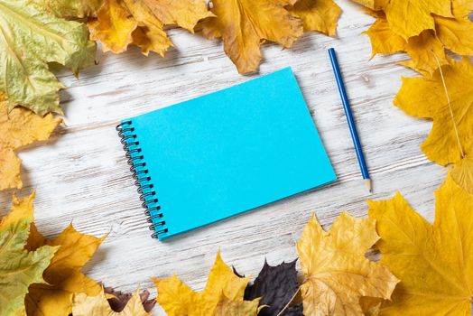 Spiral sketchbook and pen lies on vintage wooden desk with bright foliage. Flat lay with autumn leaves on white wooden surface. Blank notepad for drawing with copy space. Creative space and artwork.