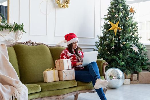 Merry Christmas and Happy New Year. Young brunette woman in santa hat sitting on green couch working on laptop in a decorated living room