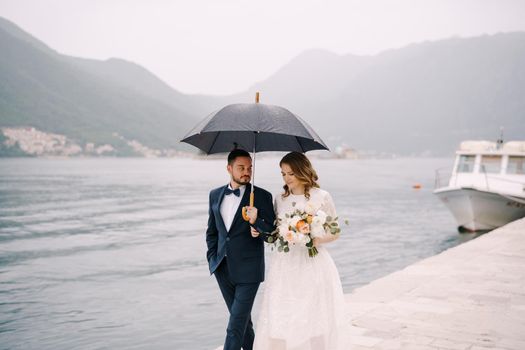 Groom and bride under an umbrella are walking along the pier. High quality photo