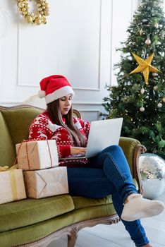 Merry Christmas and Happy New Year. Young brunette woman in santa hat sitting on green couch working on laptop in a decorated living room