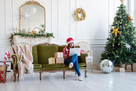 Merry Christmas and Happy New Year. Young brunette woman in santa hat sitting on green couch working on laptop in a decorated living room