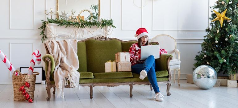 Merry Christmas and Happy New Year. Young brunette woman in santa hat sitting on green couch working on laptop in a decorated living room