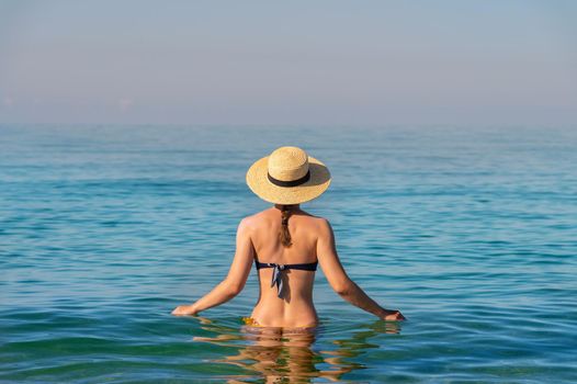 Back view of a slender caucasian young woman standing waist-deep in calm transparent sea water in a swimsuit and a straw hat.