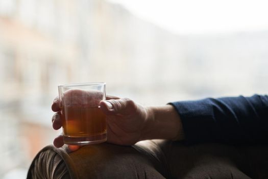 Close up shot of hand of successful business man holding glass of alcohol scotch whiskey while relaxing in a chair. Lifestyle, success, people concept