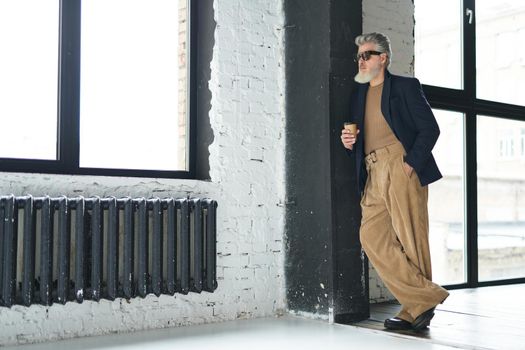 Full length shot of stylish middle aged man in business casual wear and sunglasses leaning on the wall, holding disposable coffee cup while posing in loft interior. Lifestyle, people concept