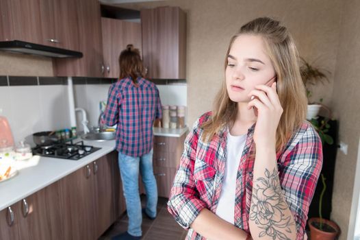 Important call. A young Caucasian woman with a serious dejected face is talking on the phone while standing in the kitchen against the background of a young husband who washes the dishes. Sorry young woman. Mortgage debts.