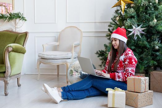 Merry Christmas and Happy New Year. Young brunette woman sitting near Christmas tree in a decorated living room working on laptop computer