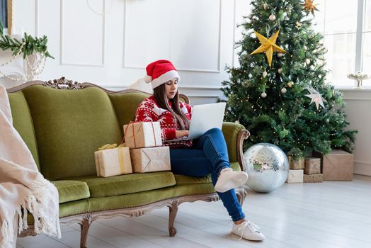 Merry Christmas and Happy New Year. Young brunette woman in santa hat sitting on green couch working on laptop in a decorated living room