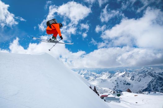 A professional skier in an orange suit jumps from a high cliff against a background of blue sky and clouds, leaving a trail of snow powder in the mountains