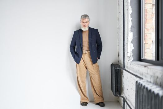 Full length shot of stylish middle aged man in business casual wear looking at camera, putting hands in pockets while posing against white background in loft interior. Lifestyle, people concept