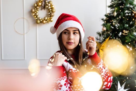 Merry Christmas and Happy New Year. Portraite of young brunette woman in santa hat and sweater holding beautiful christmas lights