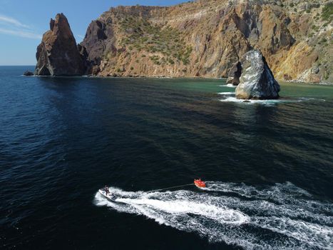 Happy people swim on air mattress behind a high speed water bike. Tourists ride on inflatable watercraft boat. Sea attraction. Water boat skating people on large Inflatable raft. Aerial copter view