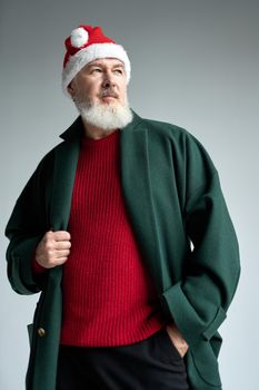 Cool middle aged man with Christmas hat wearing stylish outfit looking away, adjusting his coat while posing isolated over light gray background. Holidays concept