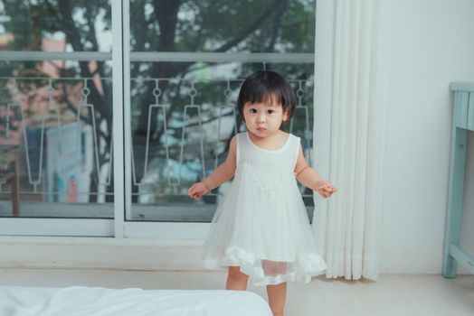 Cute toddler girl at home in white room is standing near window. 