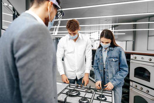 Shop assistant in hypermarket shows new model of gas stove to young couple, all wearing medical masks, close up