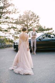 Bride walks along the road to groom standing near the convertible. High quality photo