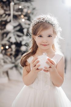 young caucasian girl with long fair hair and pretty face in white dress poses for the camera with christmas toy in her hands