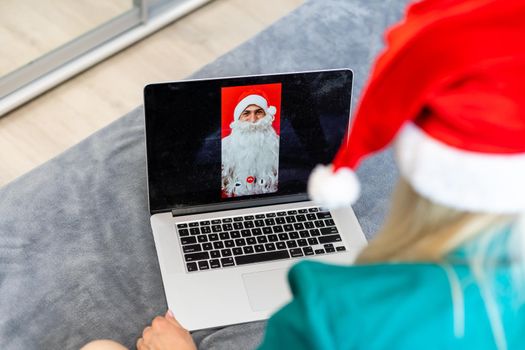 Woman in a red Santa hat communicates in a Christmas video conference. Girl celebrates Christmas at home during the pandemic. Communication with loved ones via the Internet. Online new year greetings