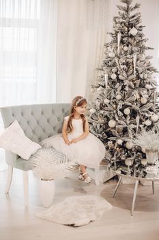 Stock photo of pretty little girl wearing head accessory and white dress with festive shoes sitting on modern couch next to beautifully decorated Christmas tree.