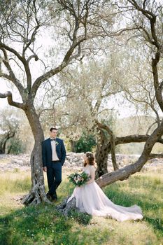 Bride sits on a tree trunk with a bouquet of flowers next to the standing groom. High quality photo