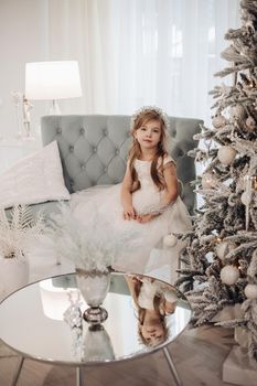 Beautiful little girl in festive head accessory and white dress sitting on grey modern couch and holding white branch looking at camera calmly. Mirrored coffee table with vase and Christmas tree.