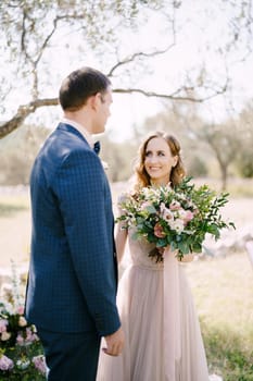 Groom stands with a smiling bride with a bouquet of flowers in a green grove. High quality photo