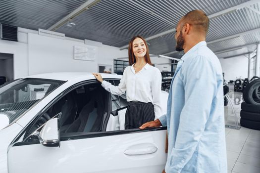 Young attractive caucasian woman salesperson in car showroom showing a car to her male African American client