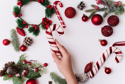 A female hand with a white manicure holds a sugar cane on a white background with Christmas branches and red decorations