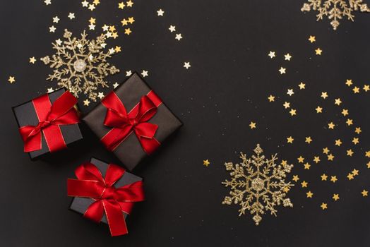 Black gift boxes with a red ribbon on a gold confetti background. Elegant Christmas background with modern golden Christmas decorations, balls, gift boxes, confetti stars on dark black background. Flat design, top view, copy space