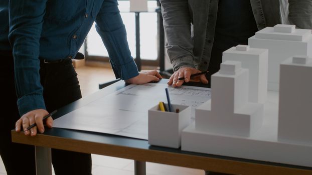 Close up of colleagues working on urban design with blueprints and building model. Team of people doing teamwork on construction layout, designing architectural development project.