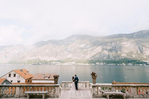 Bride hugs groom on the stone terrace against the backdrop of the bay and mountains. High quality photo