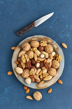 Mix of nuts on a plate - walnut, almonds, pecans, macadamia. Healthy vegan food. Clean eating, balanced diet. Blue bright table, knife for opening shell. Top view, vertical