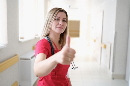 Beautiful female nurse in hospital corridor showing thumbs up, success gesture. Confident hospice worker