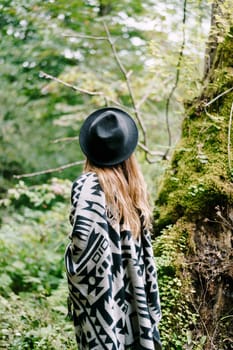 Woman in a hat and cape stands near a tree covered with moss in the park. Back view. High quality photo