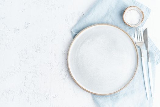 Clean empty white ceramic plate, fork and knife on white stone table, copy space, mock up, top view. Concept for menu with utensil